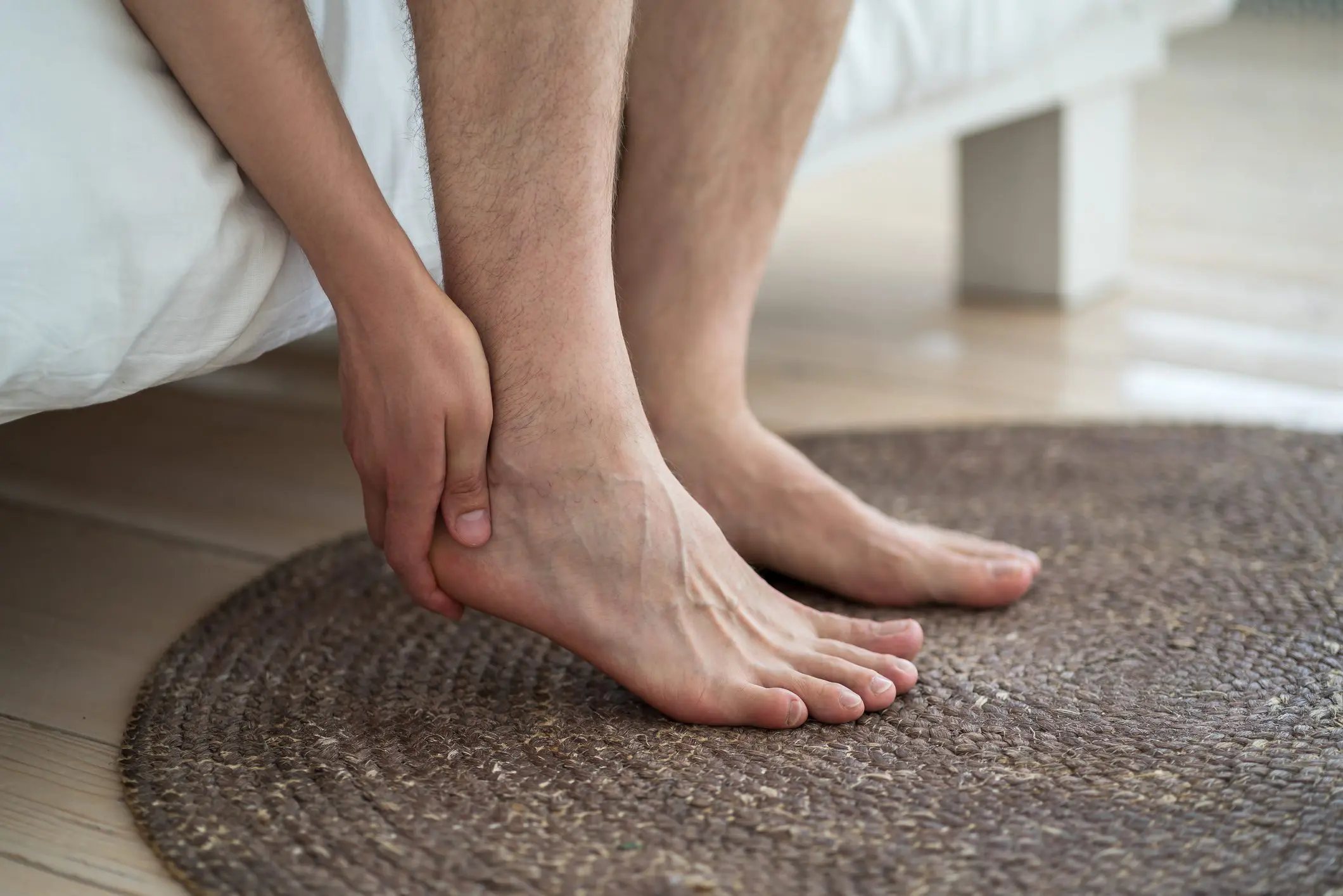 Foot pain, corns and calluses, dry skin on legs, man suffering from feet ache, podiatry concept Person sitting on bed holding their painful ankle, illustrating foot and ankle injury care and treatment services in Marion, IA.